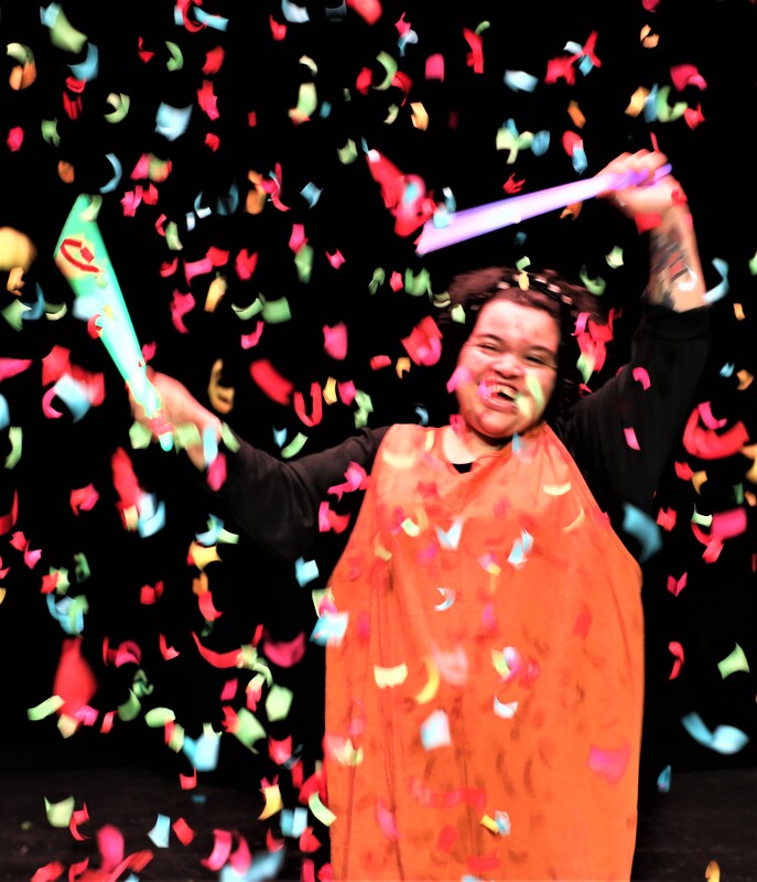 ice is a young woman of mixed heritage. She has black curly hair and is wearing a black top and orange full length playsuit. She is dancing with glow sticks under confetti on a stage. 